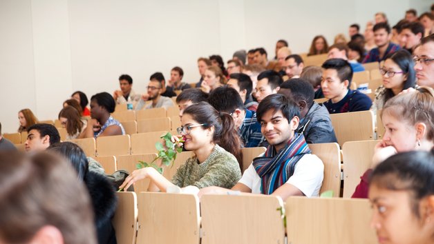 Studierende sitzen im Hörsaal, eine Person lächelt in die Kamera